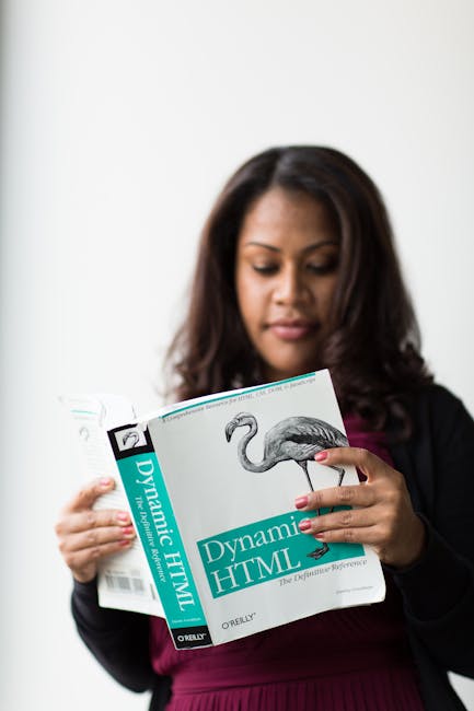 A woman with medium-length, dark brown hair wearing a black blazer and dark pink top is holding a thick book titled 'Dynamic HTML' with a flamingo illustration on the cover. She is standing on a plain